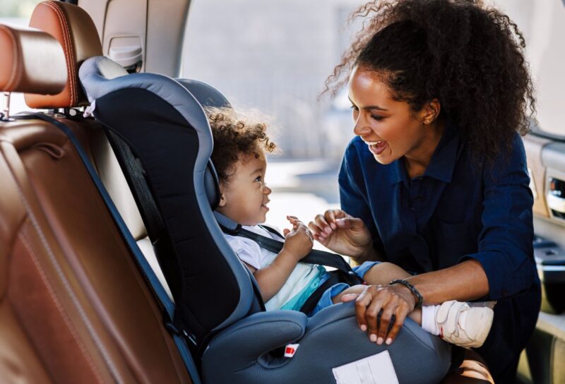 Une femme souriante attache son enfant dans un siège auto installé à l'arrière d'une voiture.